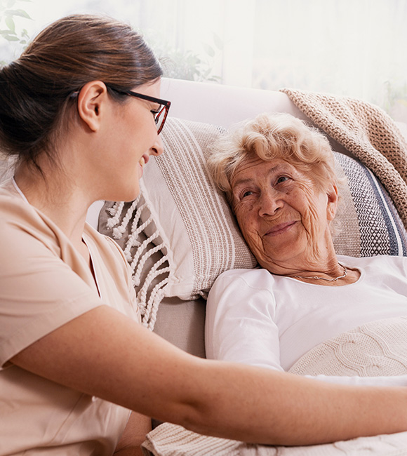 a young caregiver smiling and interacting with an elderly woman on a couch in a warm and cozy setting showcasing compassion and connection in elder care 7 days a week